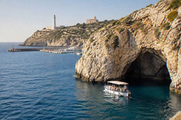 A small open passenger boat close under a tall limestone sea cliff with a sea cave opening, a few visitors on board, Santa Maria di Leuca, Puglia