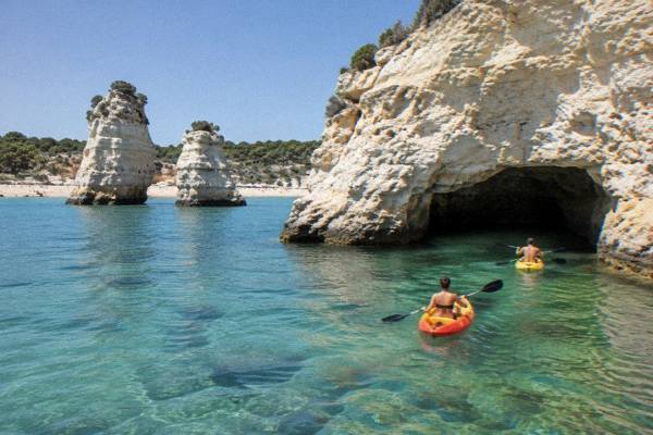 Two sea canoes paddling toward a low sea cave in a pale limestone cliff over clear green water, Torre dell'Orso, Puglia
