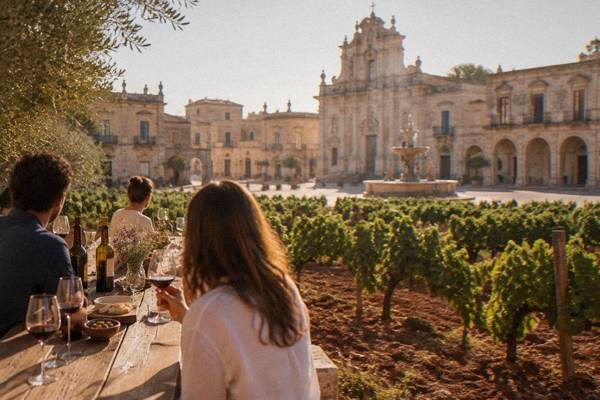 A small group tasting red wine at a long table beside a vineyard of low bush vines on red earth, Nardo, Puglia