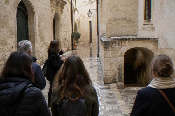 A quiet lane of the old Jewish quarter, plain pale stone walls and arched doorways, a guide talking to a small group, Museo Ebraico di Lecce, Lecce