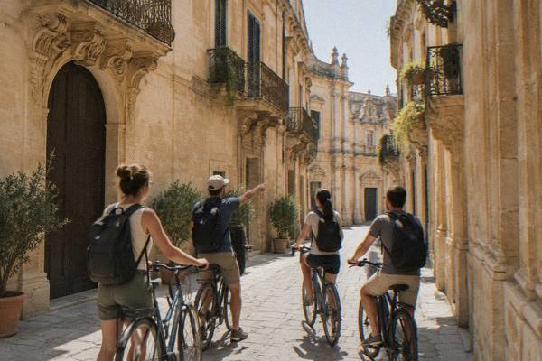 A small group on city bikes pausing in a pedestrian lane of honey-coloured stone, a guide pointing ahead, Centro Storico, Lecce