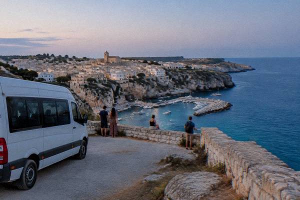 A plain white minibus parked at a clifftop viewpoint while visitors look over a deep turquoise sea, Castro, Puglia