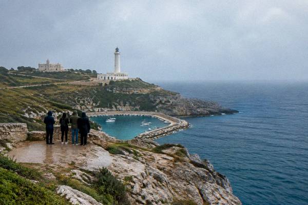 A small group of day visitors standing at the tip of a headland, a white lighthouse behind and open sea beyond, Santa Maria di Leuca, Puglia
