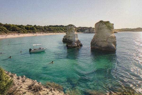 A small snorkel boat anchored near two limestone sea stacks in clear green water, a few swimmers in the sea, Torre dell'Orso, Puglia