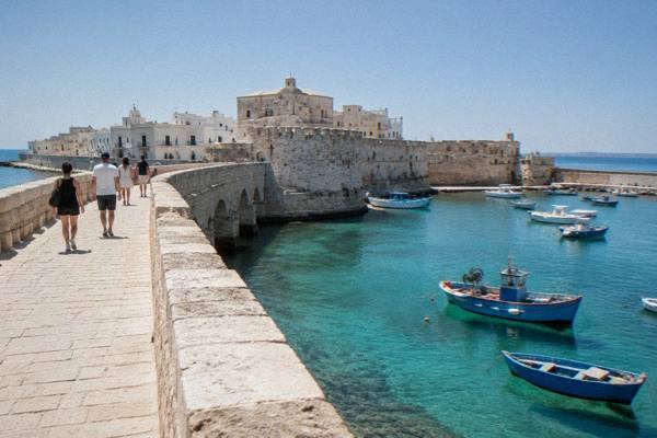 A small group walking over the stone bridge to an island old town, fishing boats in the harbour below, Gallipoli, Puglia