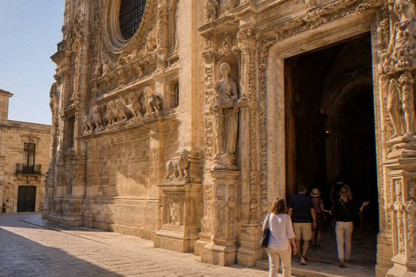A guide and a few visitors stepping through a church doorway framed by a carved stone portal, Basilica di Santa Croce, Lecce