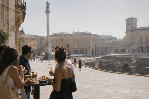 A small group sharing small plates of fried snacks and bread round a high table on a pedestrian street, glasses in hand, Piazza Sant'Oronzo, Lecce