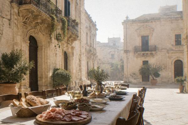 A long table in a stone courtyard laid with bread, olives, cured meats and local dishes, glasses of wine, nobody seated yet, Centro Storico, Lecce