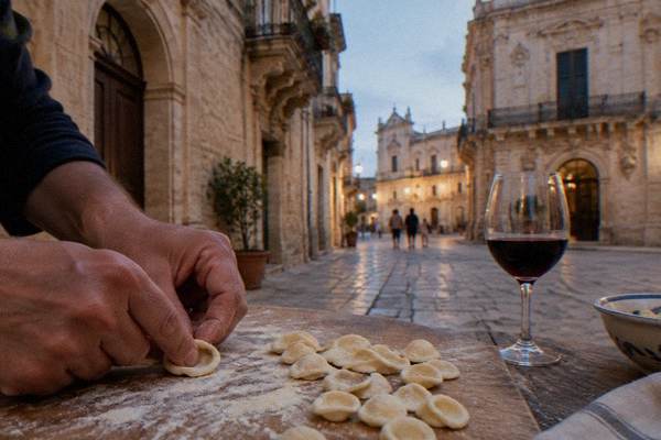 Hands pressing small orecchiette pasta shells with a thumb on a floured wooden board, a glass of red wine beside it, Centro Storico, Lecce