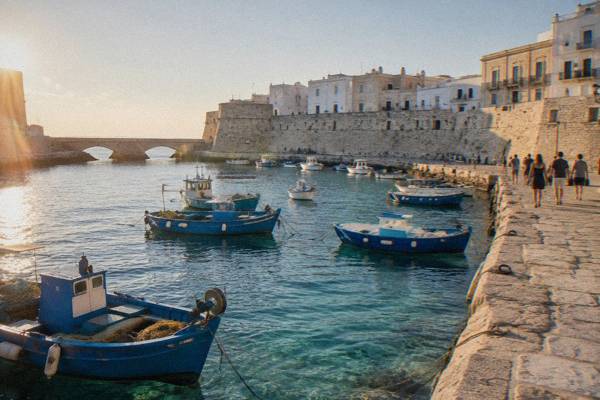 Fishing boats moored in a small harbour below an old town sea wall, a small group walking along the quay, Gallipoli, Puglia