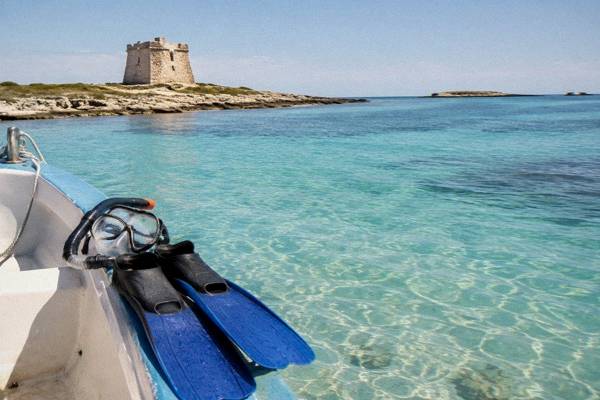 A snorkel mask and fins resting on the edge of a small boat over shallow turquoise water, a low stone tower on the shore, Porto Cesareo, Puglia