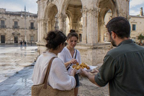 A guide handing a paper tray of fried street snacks to two visitors beside a stone arcade, Sedile, Lecce