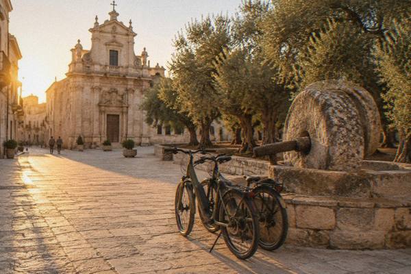 Two e-bikes parked beside an old stone olive mill, a row of ancient olive trees behind, Galatina, Puglia