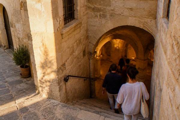 A small group stooping down a narrow stone stairway into vaulted underground rooms of honey limestone beneath an old building, Museo Ebraico di Lecce, Lecce