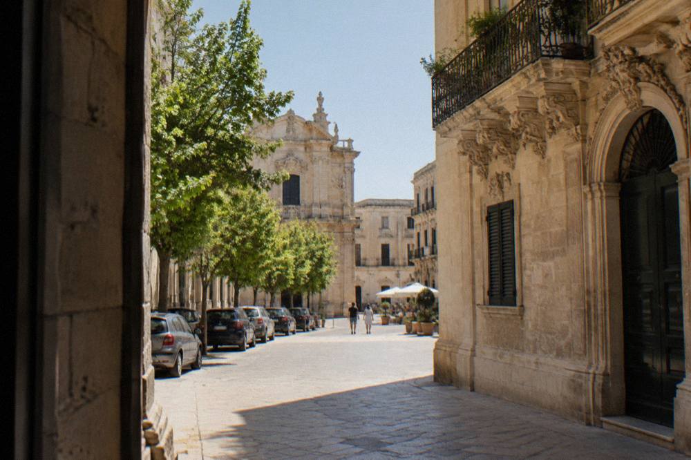 Cars parked along a tree-lined street outside a walled historic centre in southern Italy