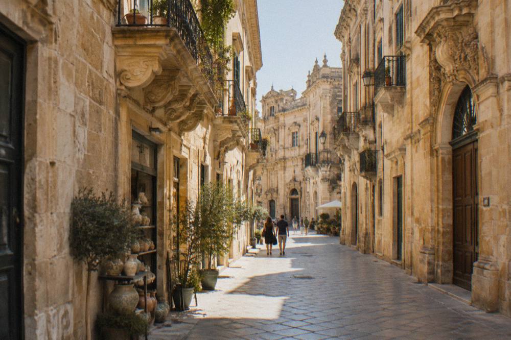 A narrow street in Lecce's old town with warm stone walls, carved balconies and small shops