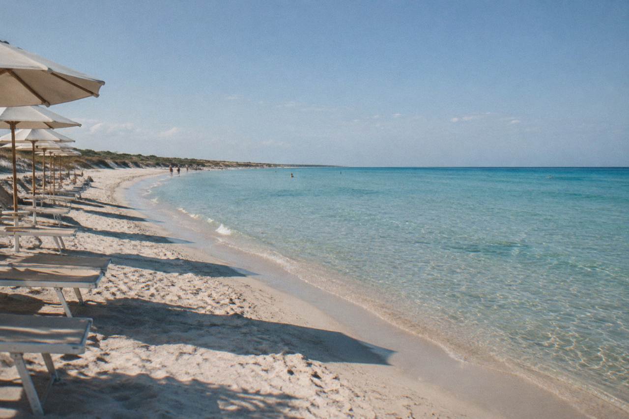 A sandy beach with clear shallow water and a row of sun umbrellas on the Salento coast