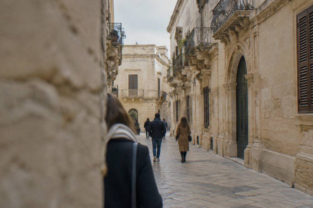 People walking down a narrow lane between pale stone buildings in Lecce