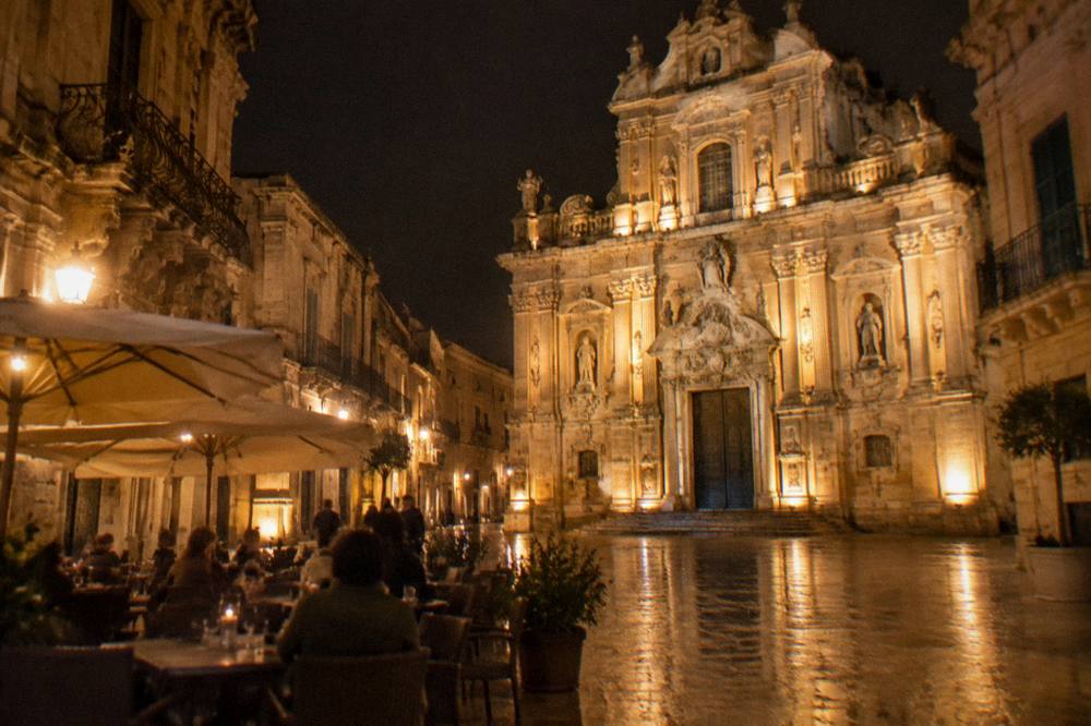 A floodlit baroque church facade in Lecce at night with people seated at outdoor tables