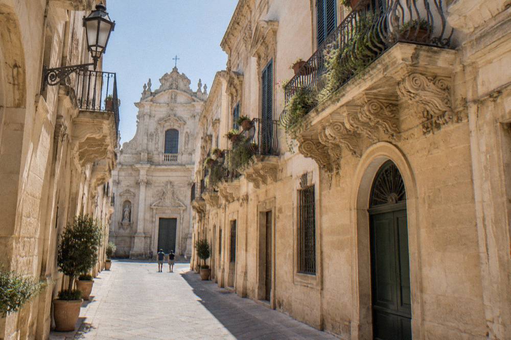A narrow old town street of pale stone buildings leading toward a church facade