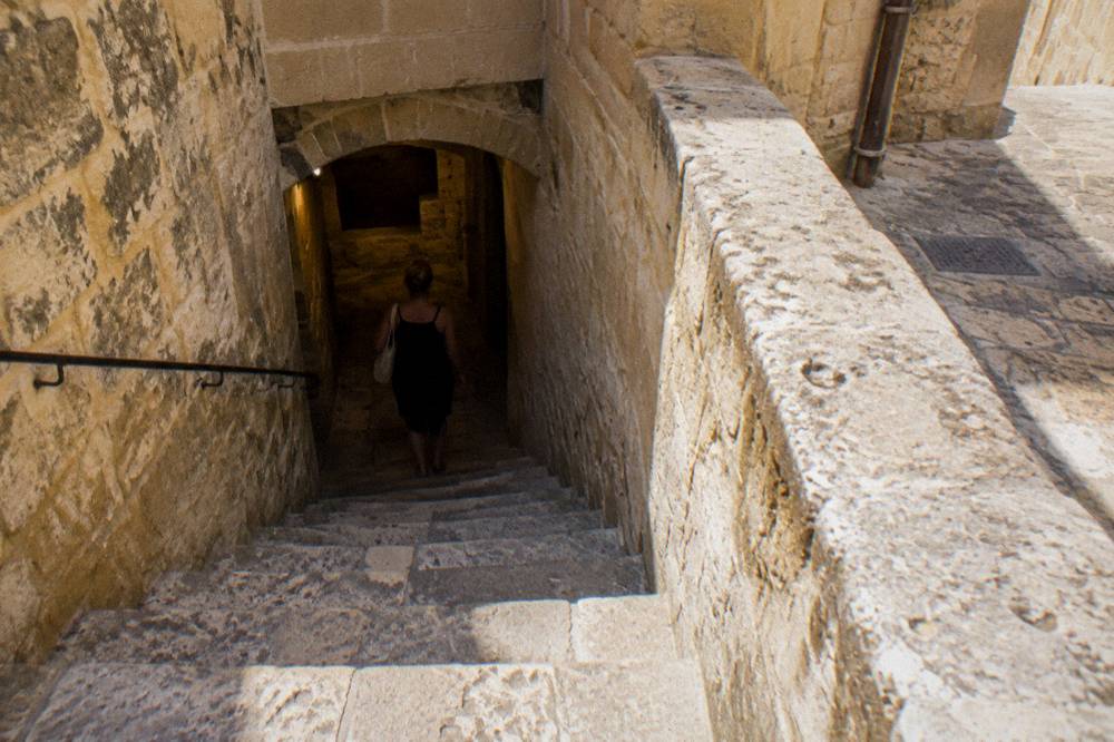 A visitor walking down worn stone steps between old walls in a dim underground passage
