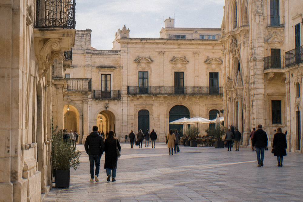A sunlit Lecce square with pale baroque buildings and people walking at dusk