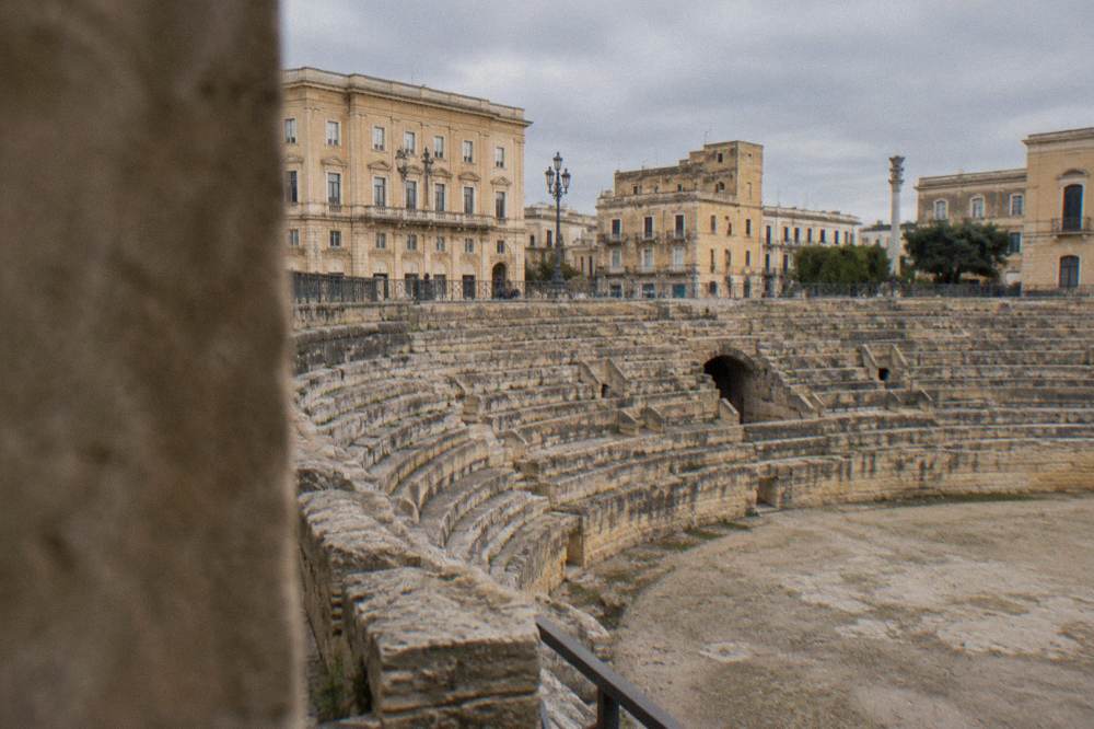 Rows of ancient stone seating descending into the excavated half of the Roman amphitheatre with modern buildings behind