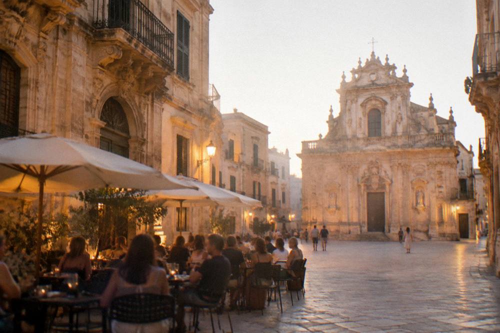 People seated at outdoor cafe tables in the square at dusk with warm street lighting on old stone buildings
