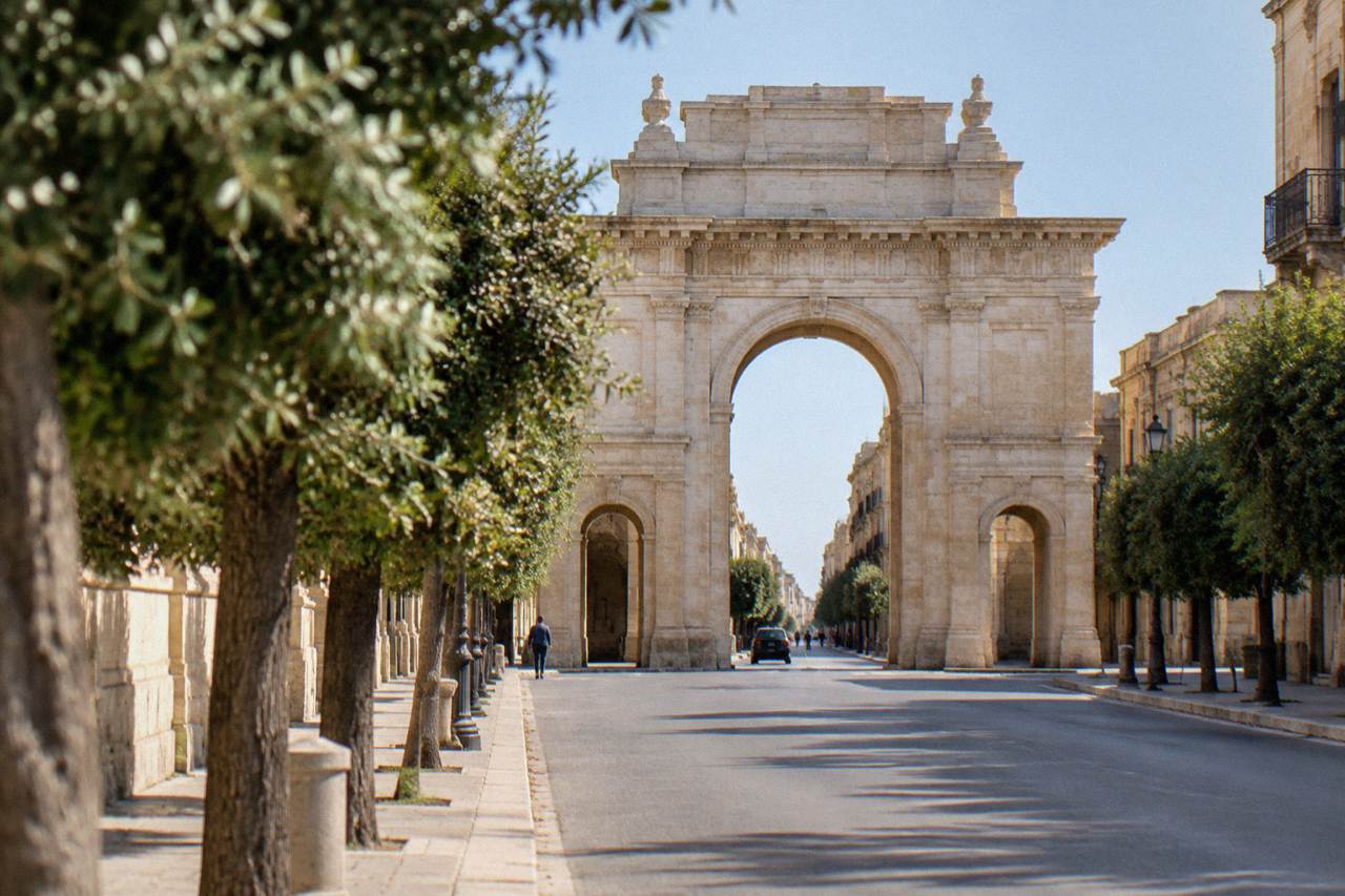 A tall stone triumphal arch with three openings standing at the end of a tree-lined city street