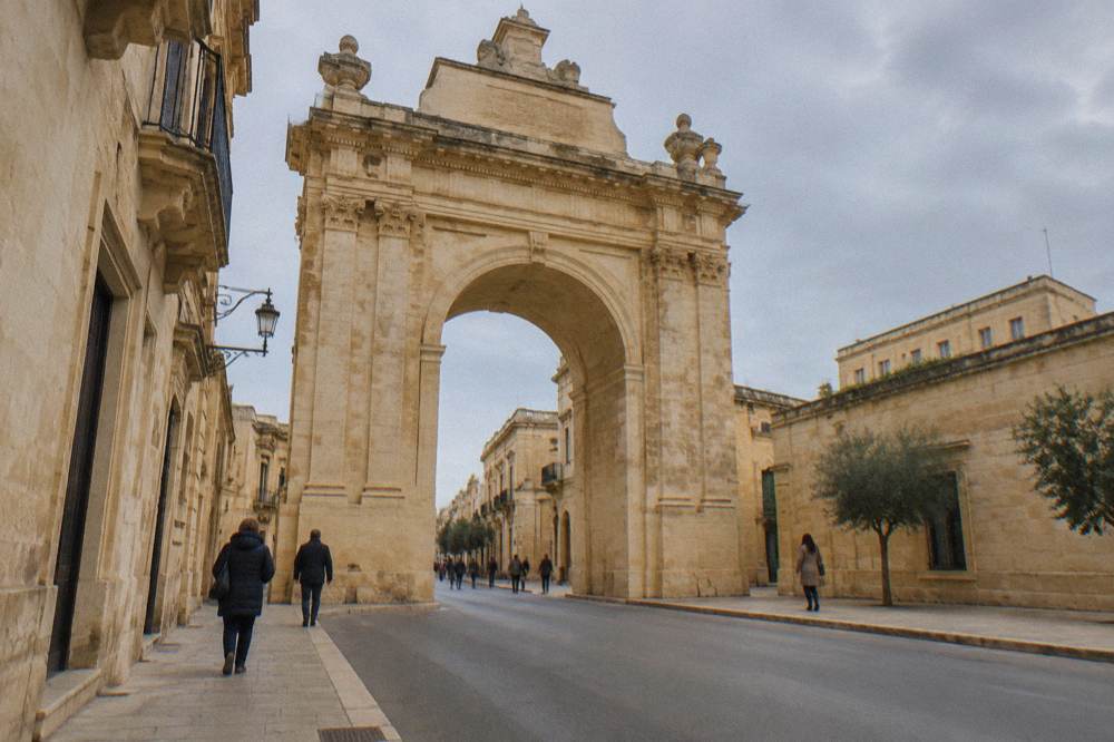 Pedestrians walking past a monumental limestone arch on a quiet morning street