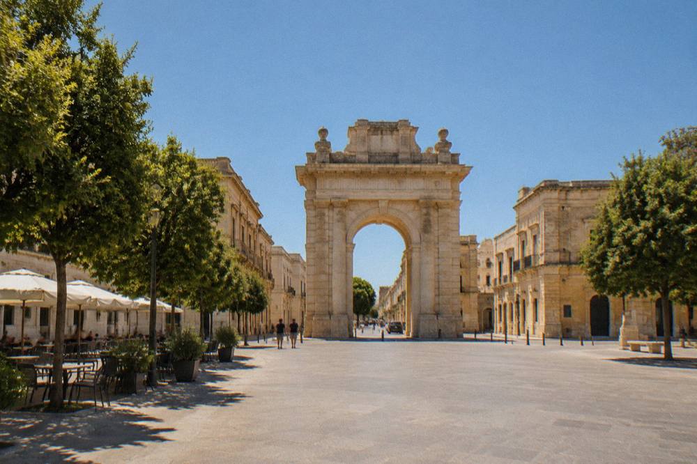 A wide city square with a stone arch at one end and cafe tables under trees