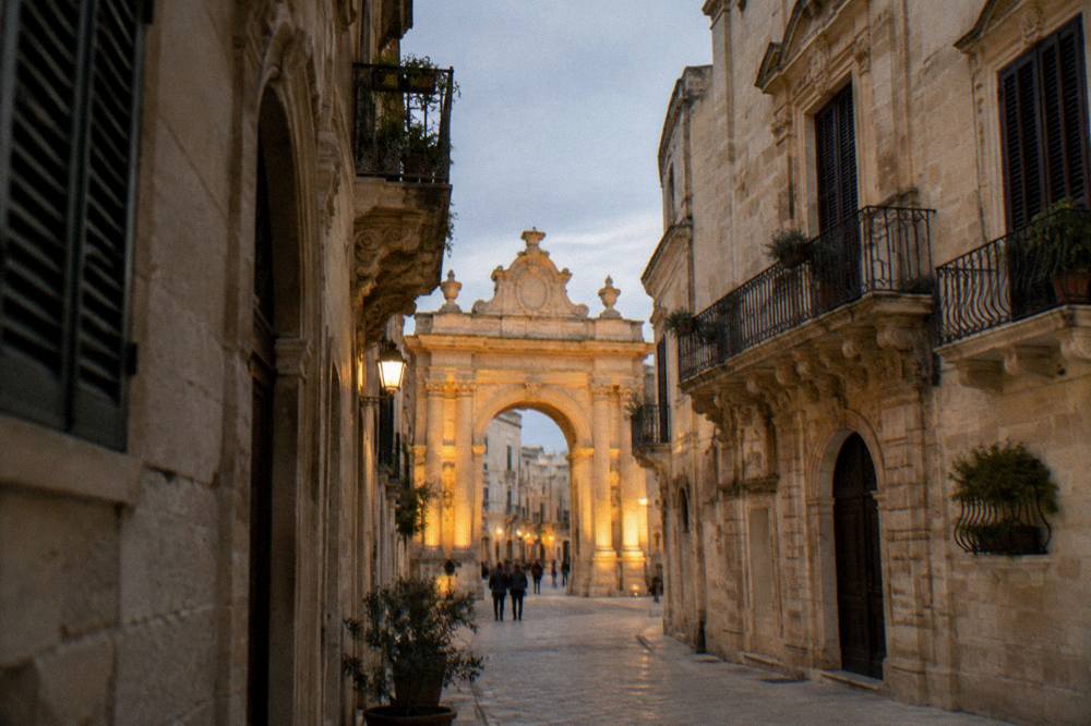 Evening view of a lit triumphal arch seen down a narrow old town street