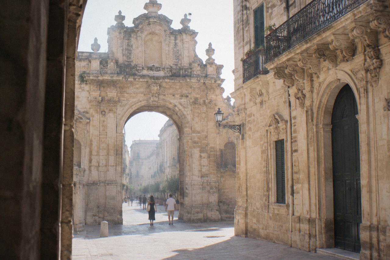 A weathered limestone city gate in Lecce with a crowded stone facade opening onto a quiet street