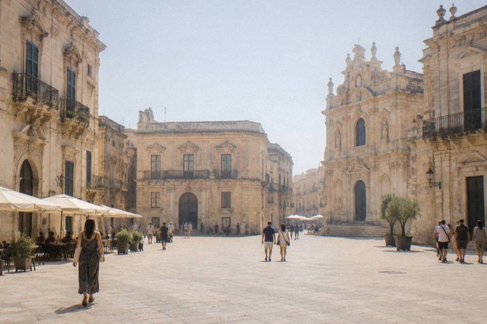 A sunlit square in Lecce with baroque limestone buildings and people walking