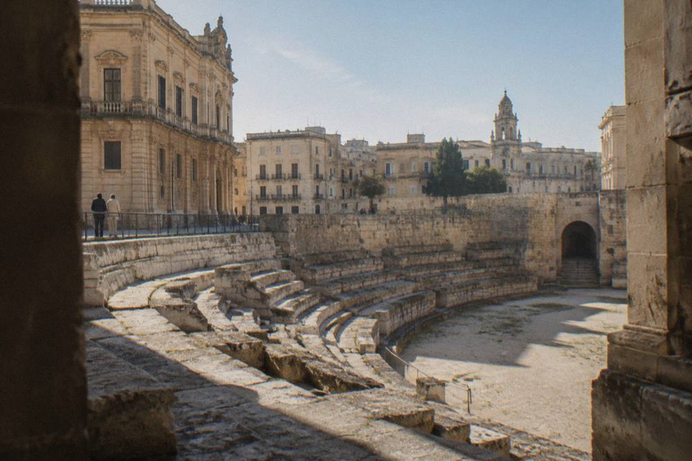 Rows of weathered limestone seating descending toward the floor of the Roman arena in Lecce