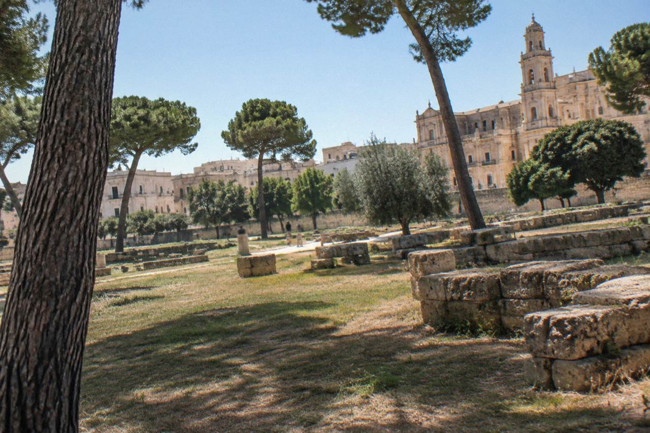 Open grassy archaeological park with low stone ruins and pine trees on the edge of Lecce