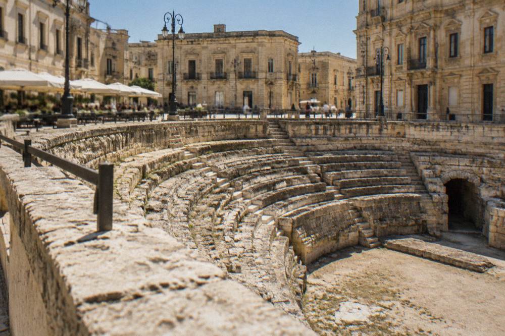 Curved rows of an ancient amphitheatre seen from a sunlit city square lined with cafes