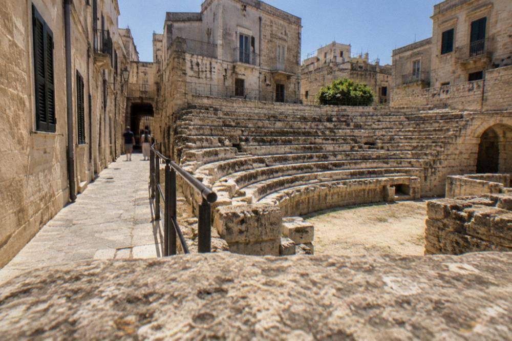 Narrow old-town lane with pale stone walls and shutters leading toward an archaeological site entrance