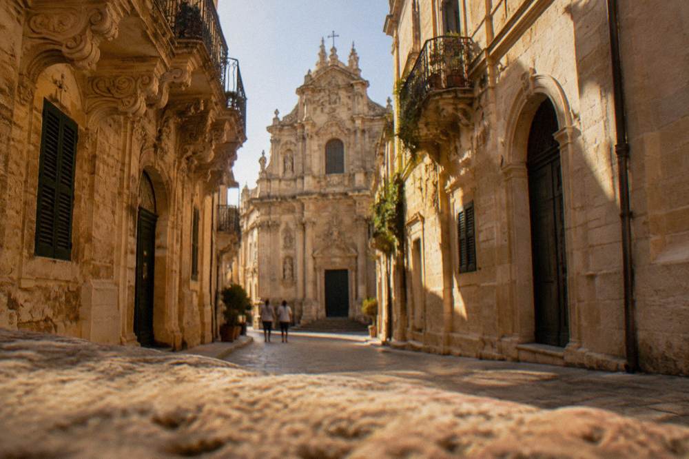 Sunlit baroque church facade with ornate carved ornament seen down a narrow Lecce street