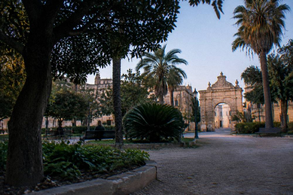 Stone city gate and garden trees seen from a quiet square at the edge of a historic centre