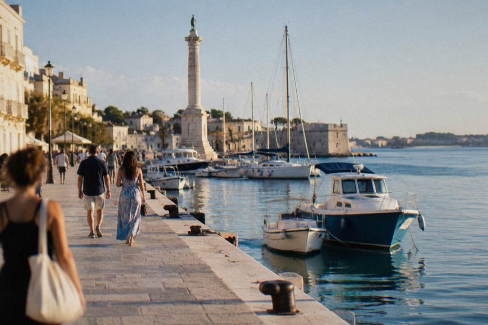 People strolling along a wide seafront promenade beside moored boats in the evening