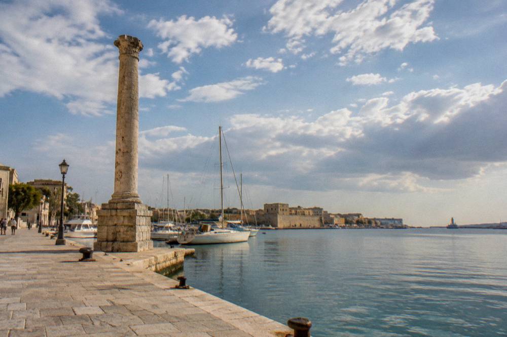 A tall ancient stone column standing on a quay beside calm sea water