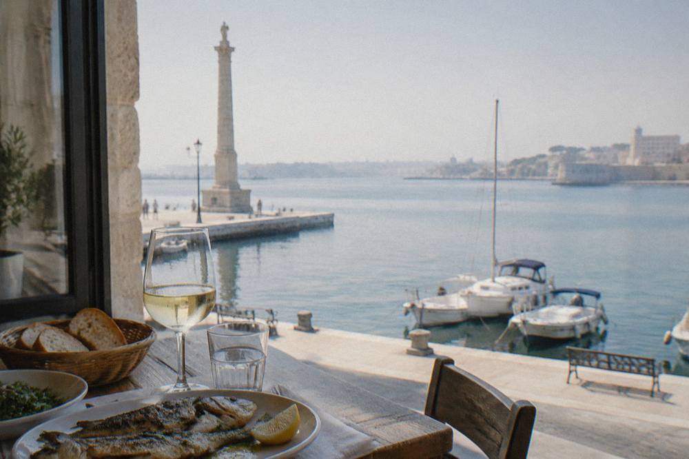 A table with grilled fish, bread and white wine beside a window overlooking a harbour