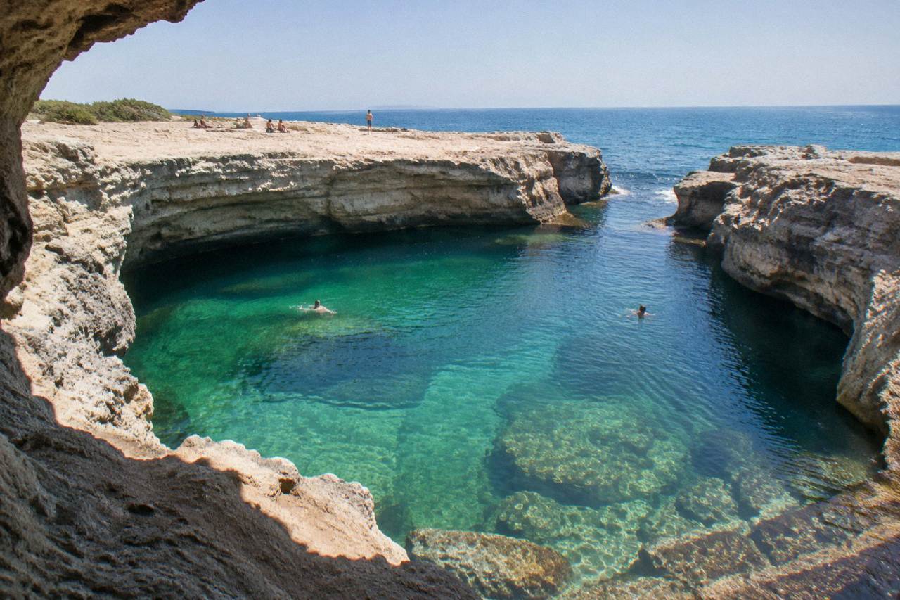 A large oval limestone sinkhole pool opening onto the sea, with rocky cliffs and clear green water