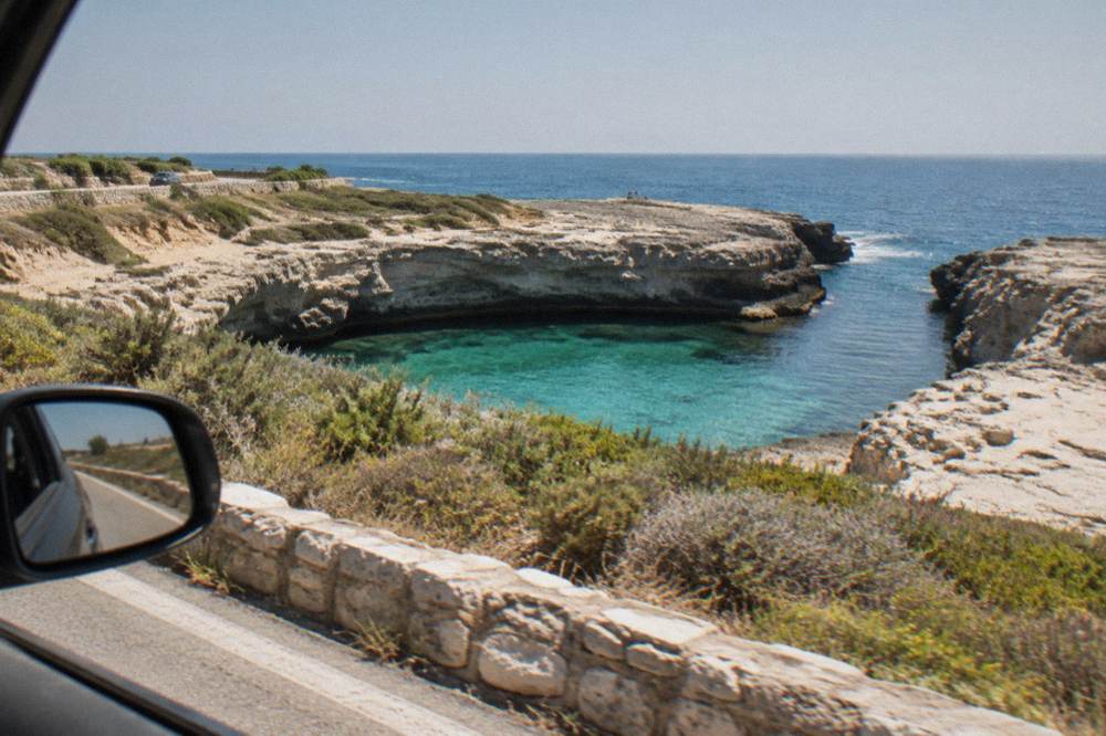 A coastal road with low Mediterranean scrub and the sea beyond, seen from a car window