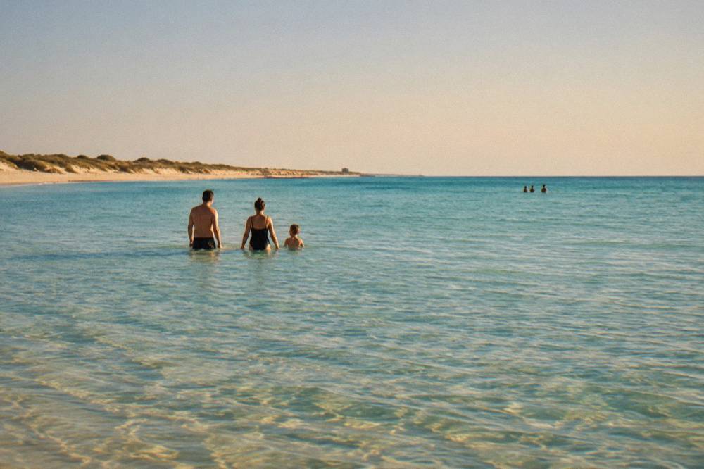 Shallow clear water over pale sand where a family wades far from the Pescoluse shoreline