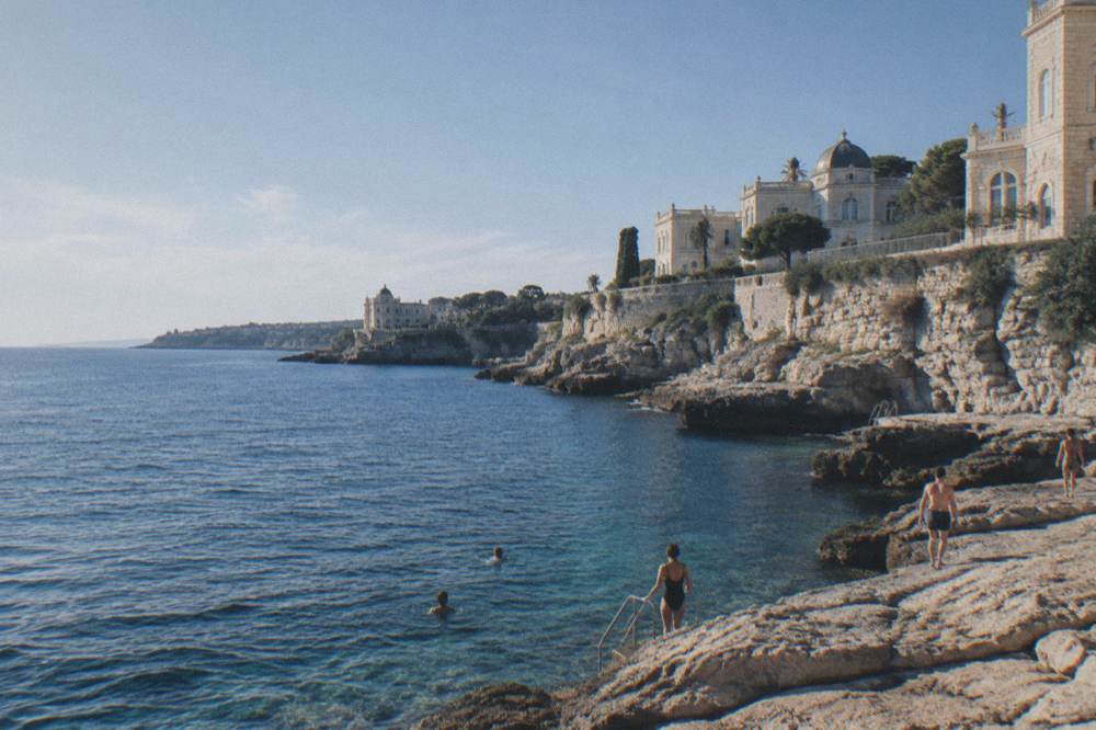 Swimmers entering clear water from flat rocks below a cliff wall