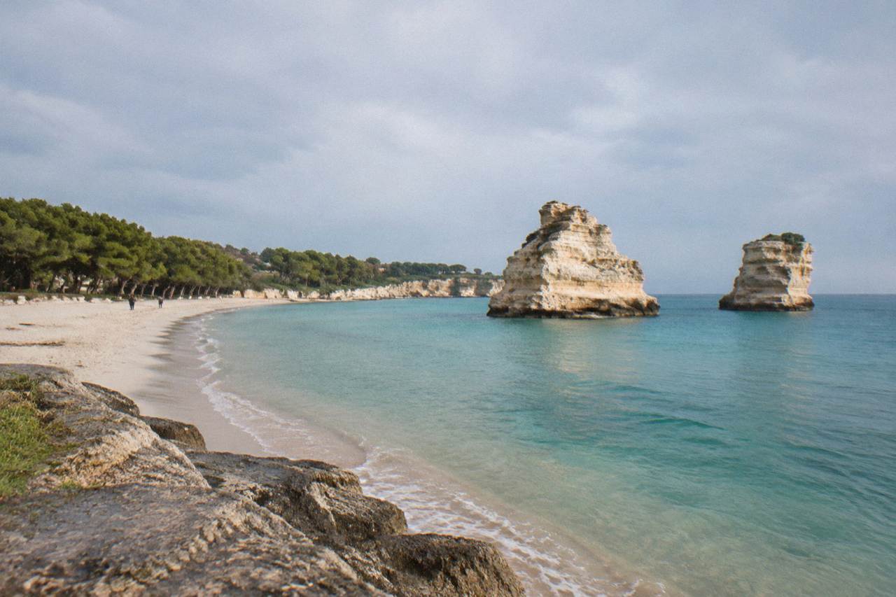 A wide sandy beach backed by pine trees, with two pale rock stacks rising from turquoise sea in the distance