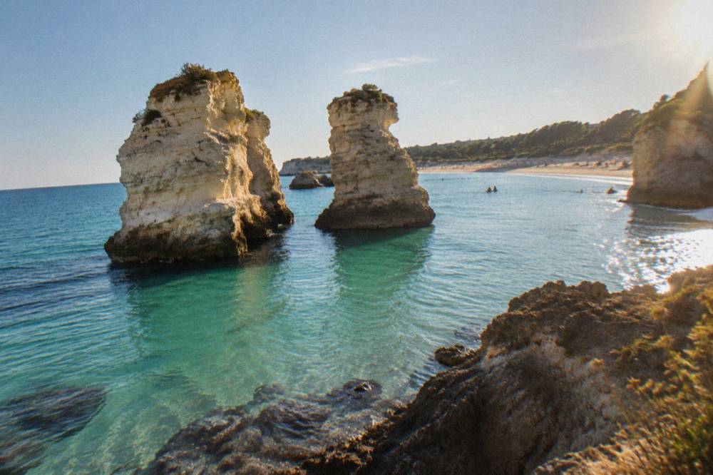 Two pale rock stacks standing close together in clear turquoise water near a sandy shore