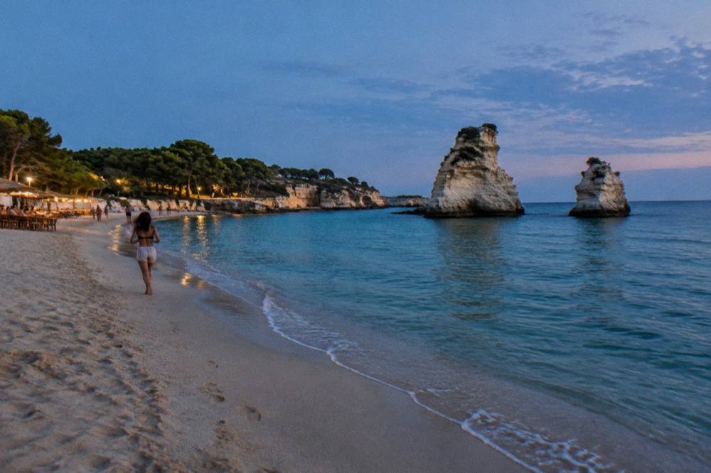 A sandy bay at dusk with pine trees, two pale limestone sea stacks in the water and a few lit beach bar tables